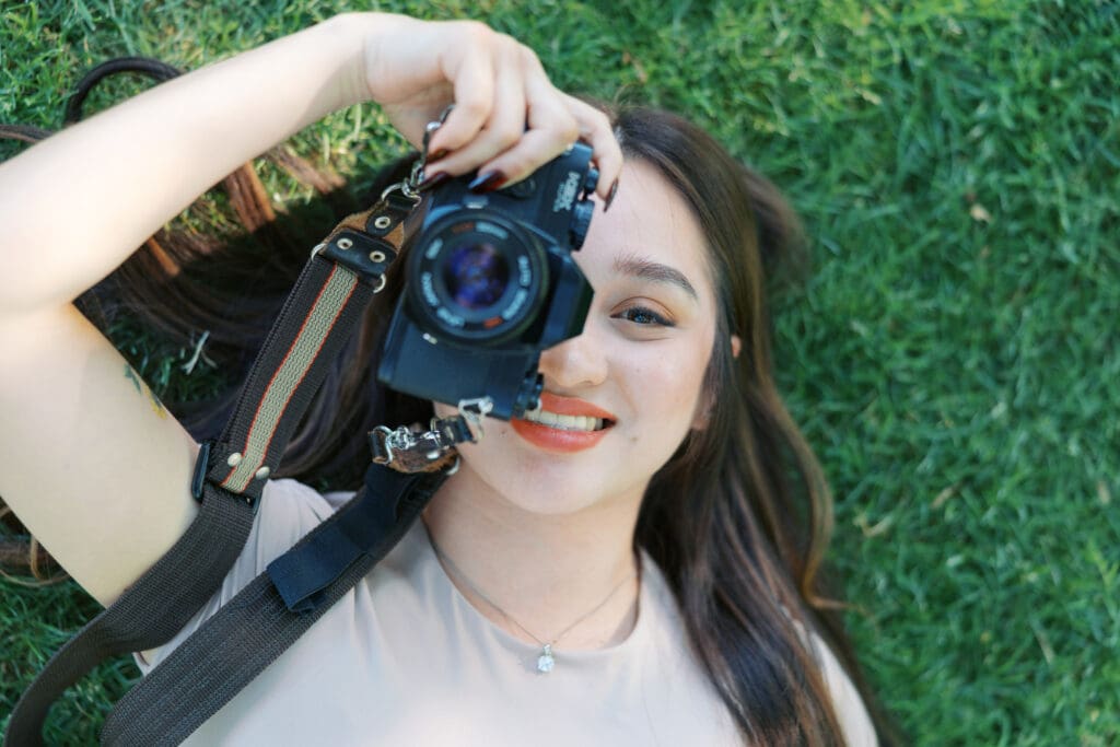 Creative entrepreneur lying on grass while holding a camera and smiling during an outdoor photoshoot.