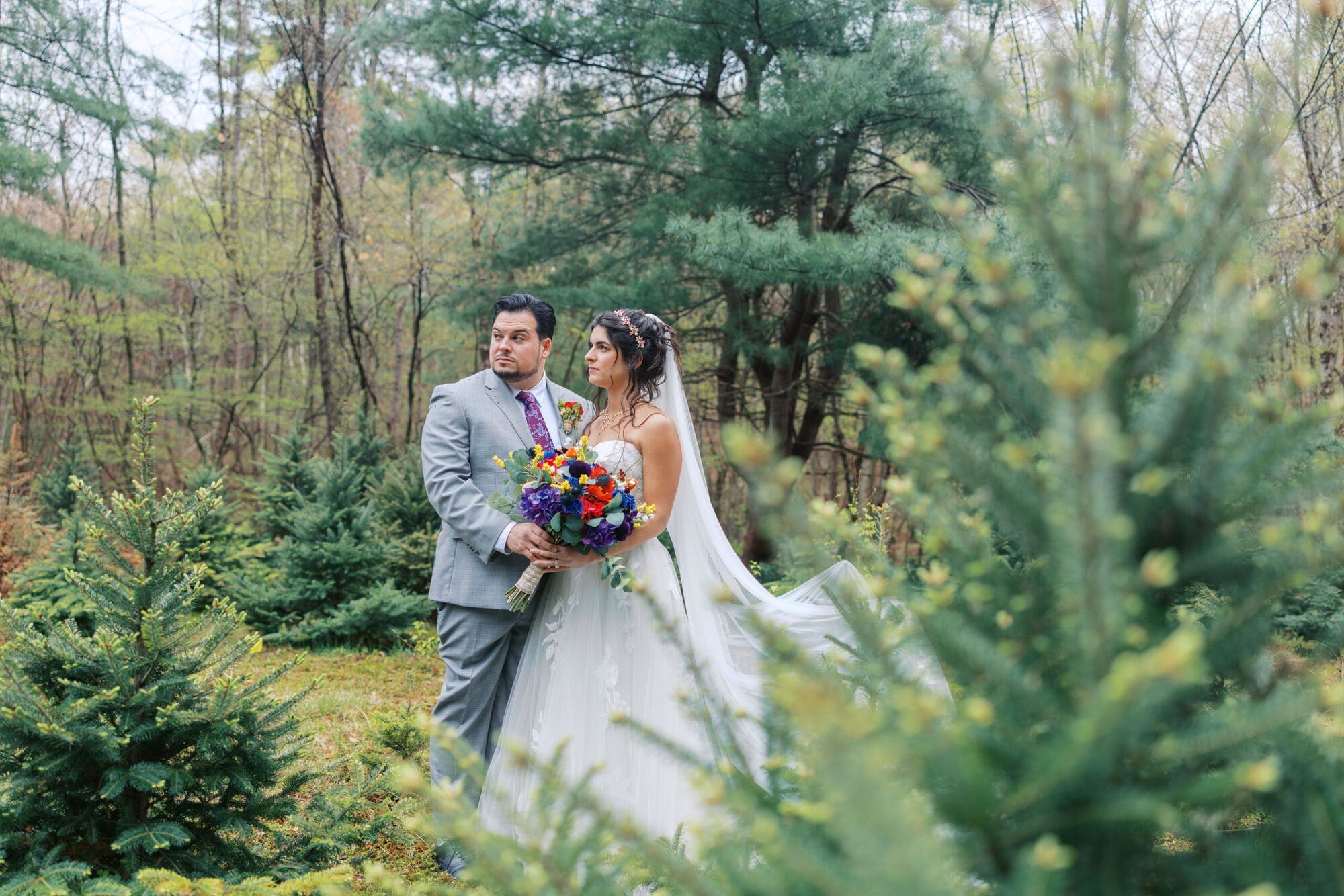Bride and groom posing naturally in a forest with colorful wedding bouquet and flowing veil during an outdoor wedding portrait session.
