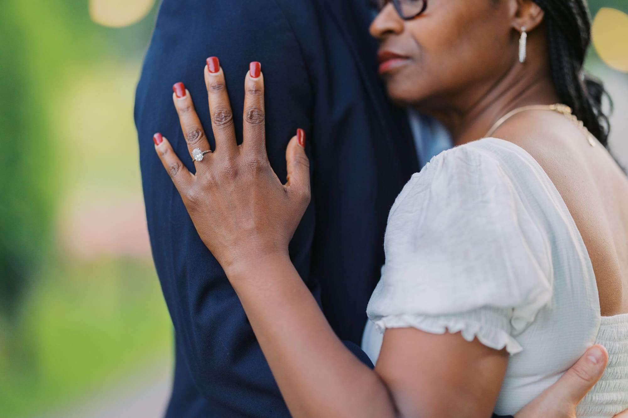 Close-up engagement photo of a couple embracing outdoors in Boston, highlighting the bride-to-be's sparkling engagement ring as she rests her hand on her partner's shoulder. The intimate composition, soft natural light, and blurred greenery create a romantic and meaningful portrait that celebrates love and connection.